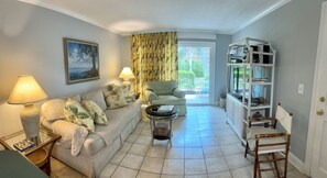 A living room with a sofa, a coffee table, a lamp, a painting, a bookshelf, and a door leading to a balcony. - E11 Peaceful 2/2 Ground Level, New Shower & Floors (St. Simons Island)