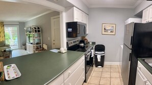 Una cocina compacta con encimera verde, armarios blancos, microondas, cubo de basura negro y refrigerador.