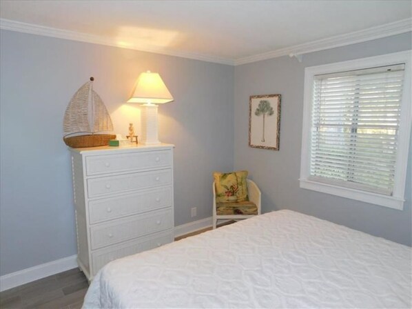 Une chambre à coucher comprenant un lit, une commode, une chaise, une lampe et une fenêtre avec des stores.