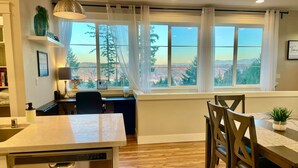 A modern kitchen with a dishwasher, wooden dining table, chairs, and a view of a mountainous landscape through large windows. - Edgewood Escape | Mt. Rainier View| EV Charger - Central (Edgewood)