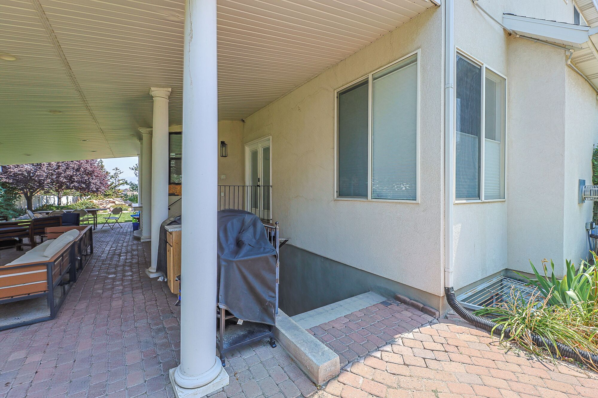 A covered patio with outdoor furniture, a grill, and a brick pathway leading to a house.