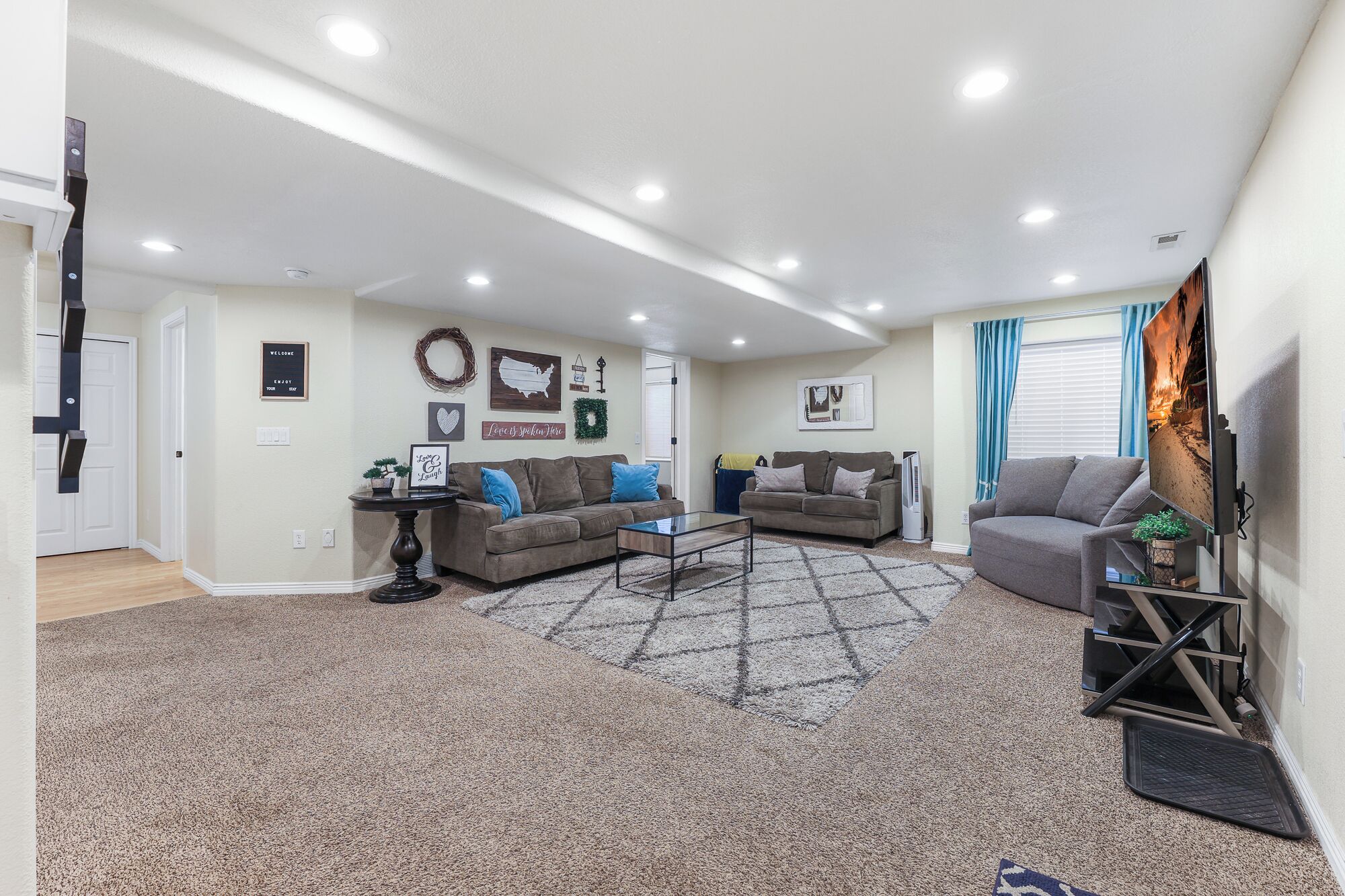 A spacious living room with two sofas, a coffee table, and a TV. There are wall decorations, a rug, and a door leading to another room.