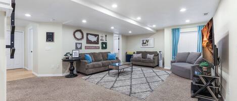 A spacious living room with two sofas, a coffee table, and a TV. There are wall decorations, a rug, and a door leading to another room.