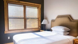 Un dormitorio con un ventanal grande, una cama con ropa blanca, una mesita de noche con lámpara y un cabecero de madera.