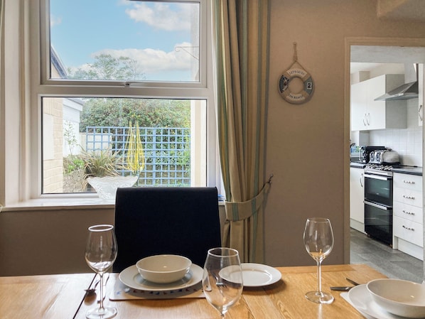 A dining area with a table set for two, a view of the garden through the window, and a kitchen visible in the background.