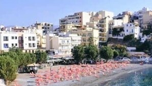 A coastal cityscape with buildings, a clear blue sea, and a beach with pink umbrellas. - Mary's Central Beachside Mansion- with large Terrace and Balcony  (Agios Nikolaos)
