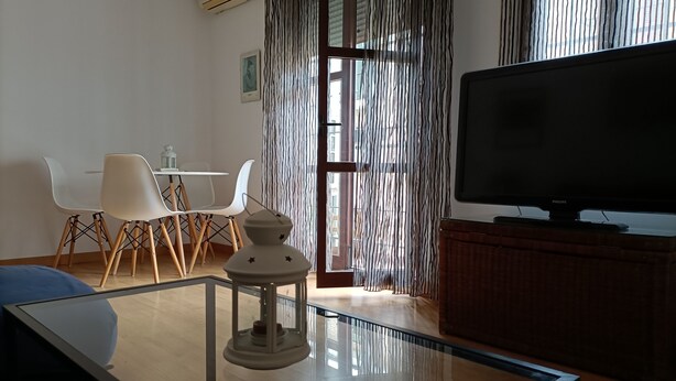 Un salon moderne avec une table basse en verre, un téléviseur à écran plat, un meuble en osier et un coin repas comprenant une table et des chaises blanches.