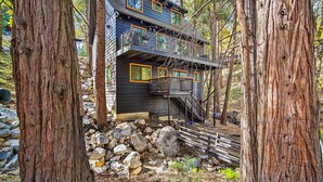 A multi-story cabin with a balcony, surrounded by large trees and a rocky ground. - Pet-Friendly Retreat w/ Game Room & Fire Pit! (Lake Arrowhead)