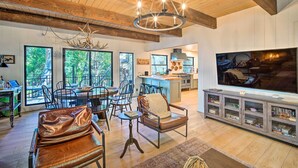 A spacious living area with a large TV, wooden ceiling beams, and a dining area with natural light from large windows. - Pet-Friendly Retreat w/ Game Room & Fire Pit! (Lake Arrowhead)