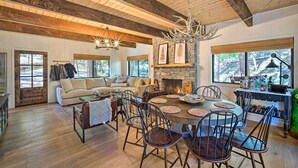 A spacious living room with a fireplace, wooden ceiling beams, and a dining area with a round table and chairs. - Pet-Friendly Retreat w/ Game Room & Fire Pit! (Lake Arrowhead)