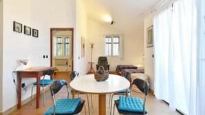 A dining area with a round table, four chairs, and a small table with a lamp. There are two windows with blinds, a door, and a wall with framed pictures. - The whole Mario's house for rent (Jelsa)