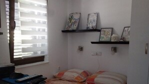 Une petite chambre à coucher, de dimensions réduites, comprenant un lit, un bureau avec un ordinateur et une télécommande, une fenêtre avec des stores et une étagère ornée d’objets décoratifs.