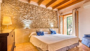 A bedroom with a stone wall, a bed with blue pillows, a wooden headboard, a nightstand with a lamp, and a view through a window. - Charming Pollensa Villa | Beautiful Views Of the Tramuntana Mountains | La. (Pollensa)