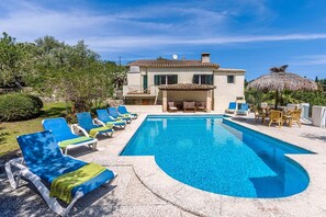 Una piscina con sillones de descanso, un edificio con terraza y una sombrilla de paja.