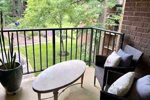 A balcony with a white table and chairs, a potted plant, and a view of a green lawn and trees.