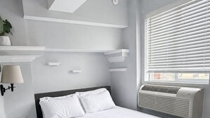 A bedroom with a bed, a wall-mounted air conditioner, and a window with blinds.