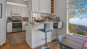 A modern kitchen with white cabinets, a microwave, and a stove. There is a bar area with stools and a view of a mountainous landscape through a large window.