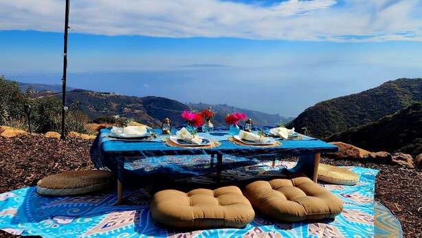 An outdoor dining setup with a table set for four, surrounded by a patterned blanket, on a rocky terrain with a view of distant hills.