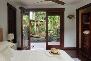Une chambre avec un lit, un chapeau et une vue sur un jardin tropical, visible à travers une porte ouverte.