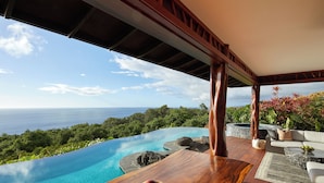 Une table en bois avec un saladier de fruits, une piscine naturelle, un jacuzzi et une vue sur l’océan.