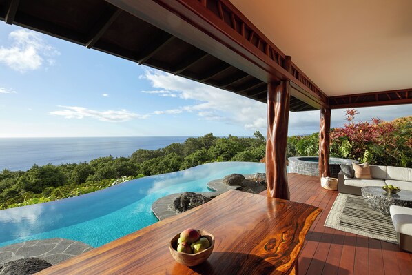 Une table en bois avec un saladier de fruits, une piscine naturelle, un jacuzzi et une vue sur l’océan.