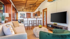 Ein modernes Wohnzimmer mit einer Couch, einem grünen Sessel, einem Couchtisch mit Obst, einem Fernseher und einer Küchenzeile mit Barhocker und einer Mikrowelle.