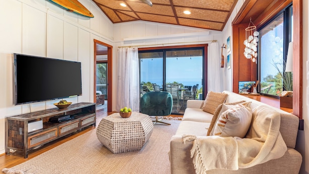 Ein Wohnzimmer mit einem großen Flachbildfernseher, einer Holzunterhaltungsecke, einem grünen Sessel, einem beigen Sofa und Blick auf den Ozean durch große Schiebegläser.