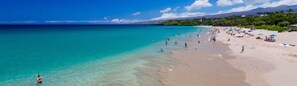 A beach with clear turquoise water, people swimming and sunbathing, and a line of trees in the background.