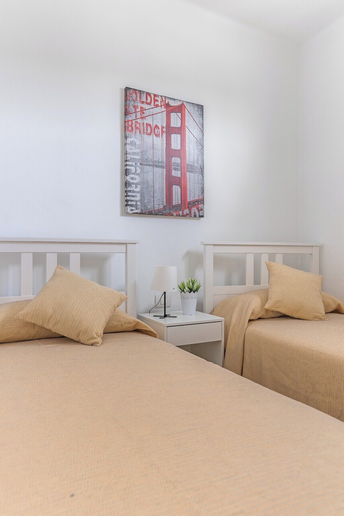 Habitación de hotel con dos camas, una mesita de noche con una lámpara y una planta, y un cuadro en la pared del Puente Golden Gate.