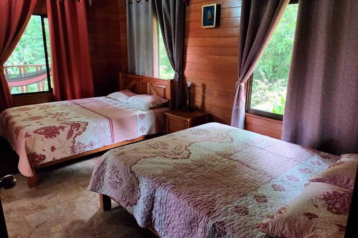 A room with two beds, wooden walls, a window with curtains, and a small table.