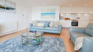 En moderne stue med en blå sofa, et sofabord i glas og et køkken med hvide skabe og et indbygget ovn.