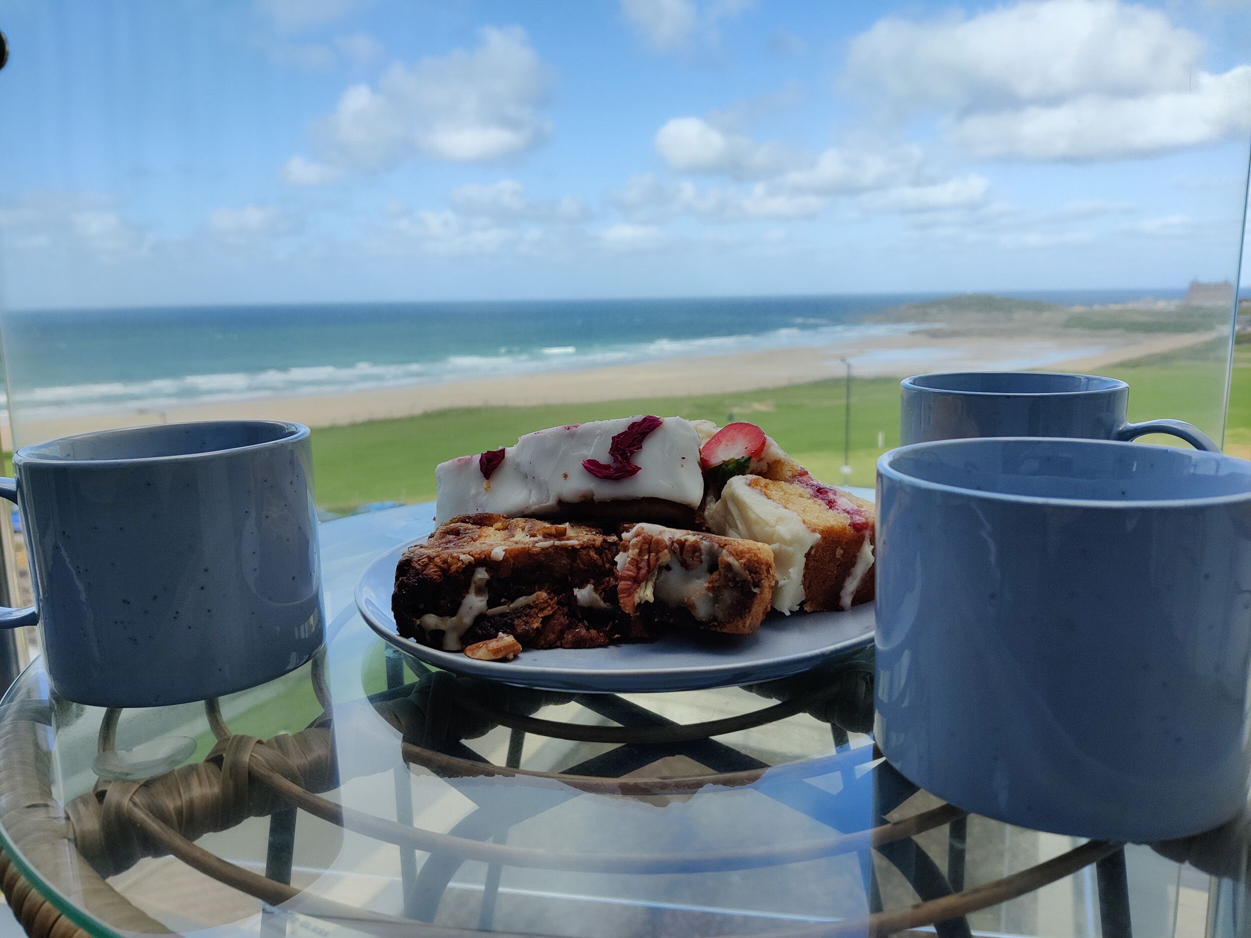 A glass table with a plate of food and two mugs, overlooking a beach and ocean.