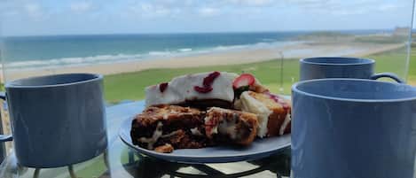 A glass table with a plate of food and two mugs, overlooking a beach and ocean.