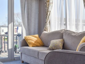 A grey sofa with two cushions, one fluffy and one with a floral pattern, in a room with large windows and curtains. - Oh I do like to be beside the seaside. (West Mersea)