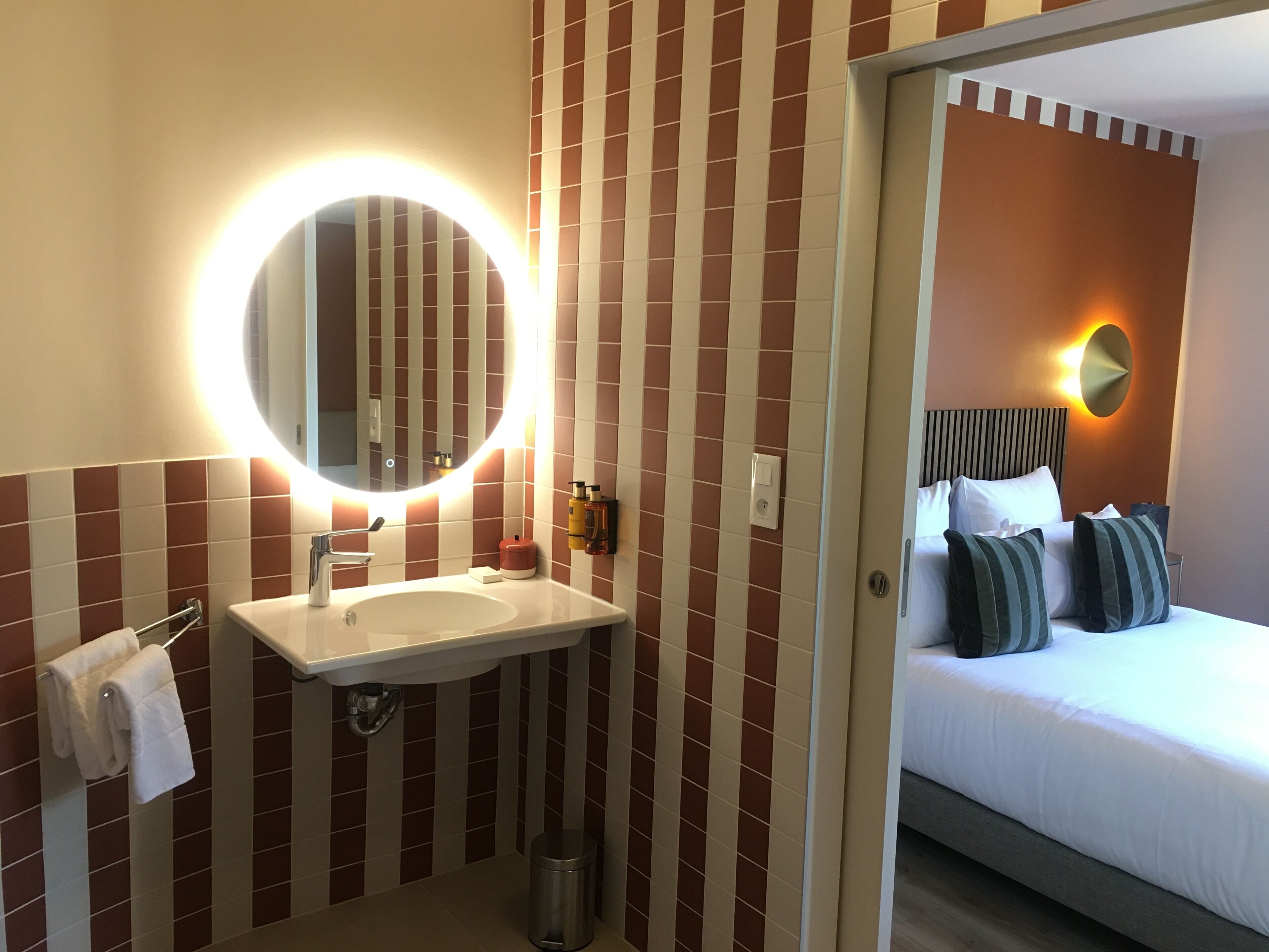 A hotel bathroom with a sink, mirror, and toilet. The room features a bed with pillows and a bedside table.