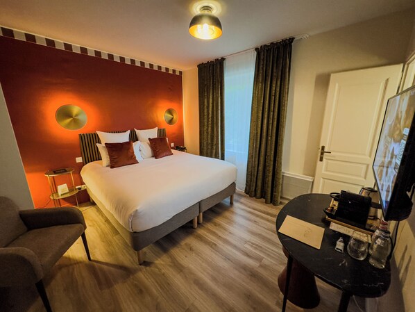 Hotellihuone, jossa on suuri sänky, yöpöytä, tuoli, työpöytä tietokoneella ja peili.
