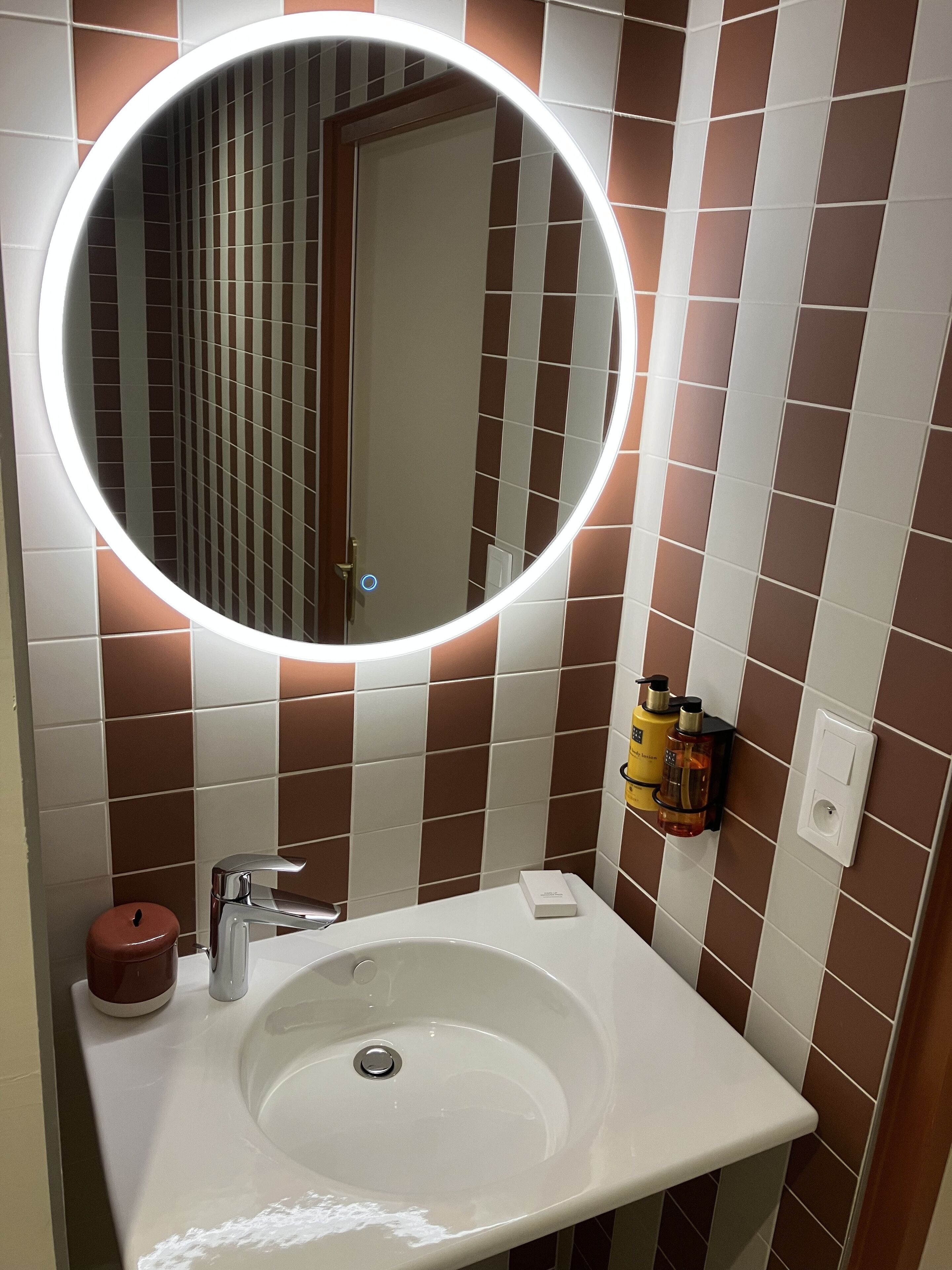 Un baño con un lavabo blanco, un espejo redondo con iluminación y baldosas marrones y blancas en la pared.