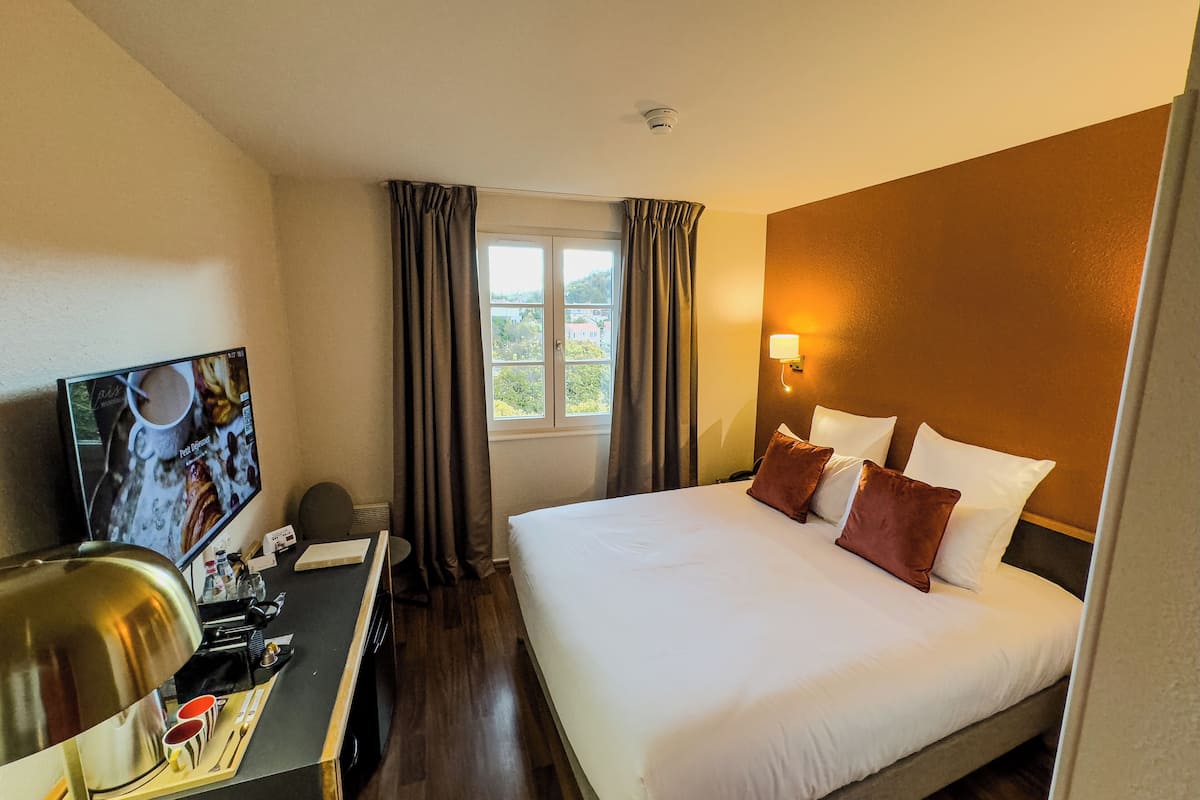 A hotel room with a bed, a desk with a lamp, a television, and a window with curtains.