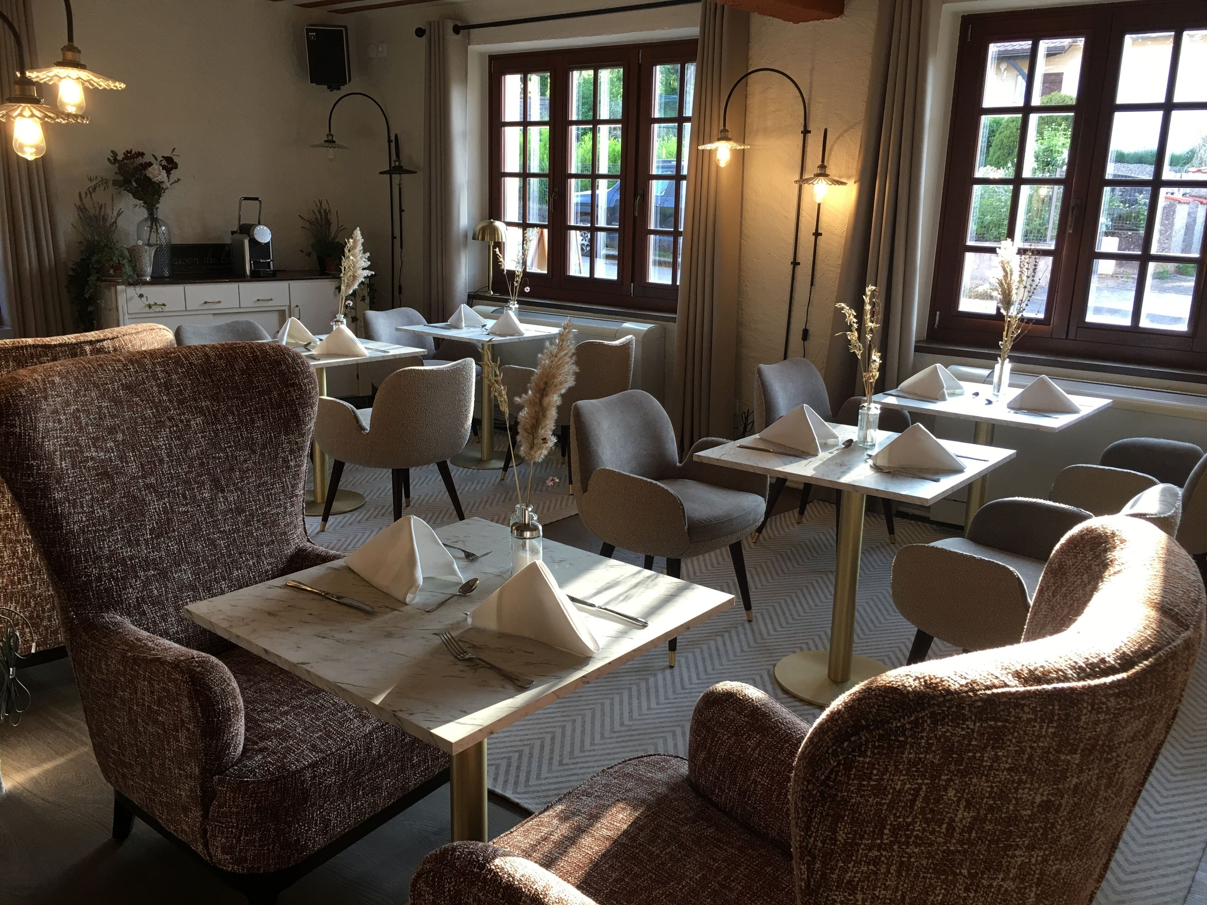 A dining area with multiple tables and chairs, some with napkins and cutlery. There are decorative plants and a window with curtains.