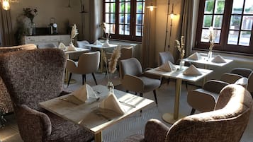 A dining area with multiple tables and chairs, some with napkins and cutlery. There are decorative plants and a window with curtains.