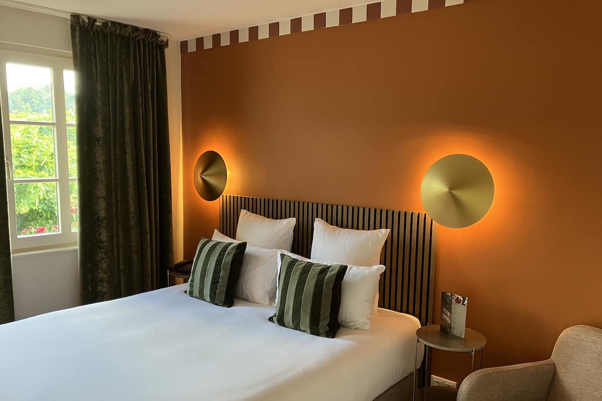 A hotel room with a large bed, two bedside lamps, a chair, and a view of greenery through the window.