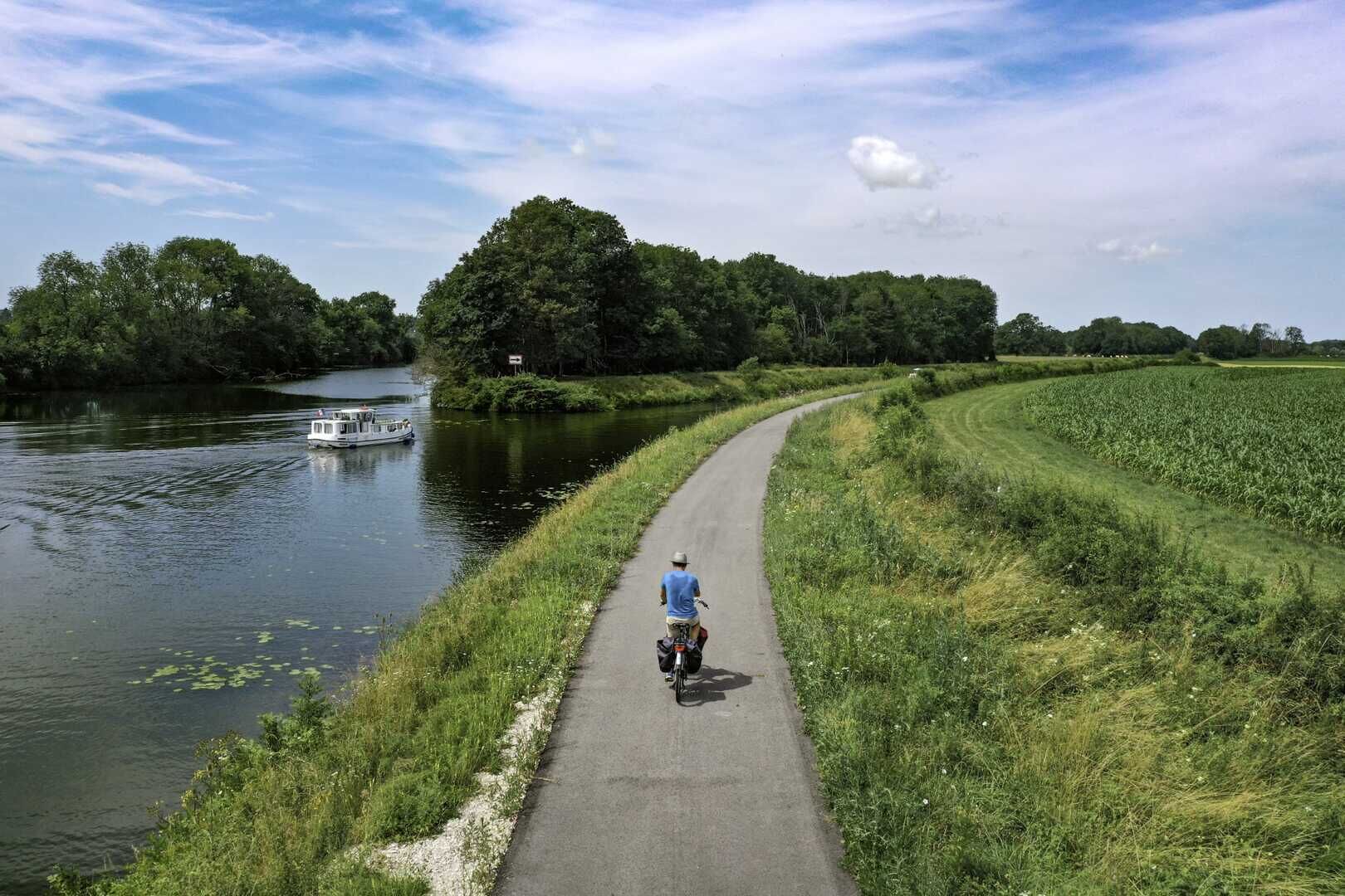 Un camino pavimentado junto a un río, con una barca en el agua y un ciclista a lo lejos.