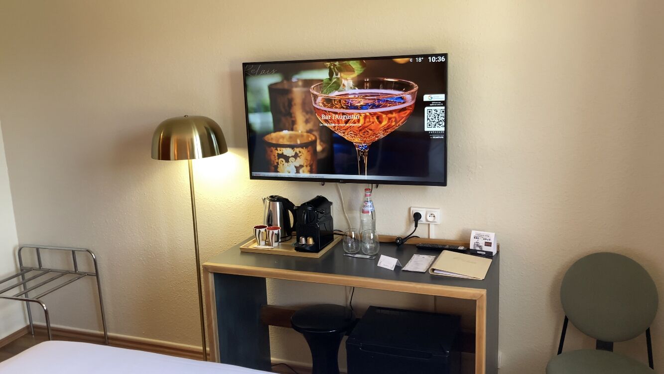 A hotel room with a TV displaying a cocktail, a desk with a coffee maker, a bottle, and a glass, and a chair.