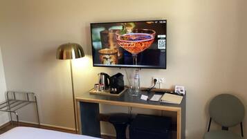 A hotel room with a TV displaying a cocktail, a desk with a coffee maker, a bottle, and a glass, and a chair.