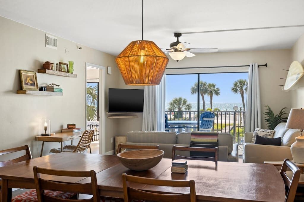 A spacious living area with a dining table, chairs, a sofa, and a balcony with a view of palm trees and the ocean.