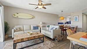 Ein geräumiges Wohnzimmer mit Deckenventilator, einem Surfbrett an der Wand, einem Holztisch und einem Essbereich mit Holztisch und Stühlen.