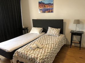 A double bed with a floral bedspread and a plain white bedspread, a nightstand with a lamp, and a painting of a cityscape on the wall.