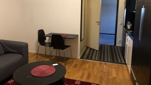 A compact living space with a sofa, a small round table, a dining table with chairs, a kitchen area with a refrigerator, and a door leading to another room. - Apartment in Årsta Stockholm 244 (Arsta)