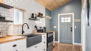 A kitchen with white cabinets, a black sink, and a wooden countertop. There is a window with blinds, a door with a sign, and a shelf with jars. - Bryce & Zion Midpoint w/ Memorable Cowboy Hot Tub (Alton)