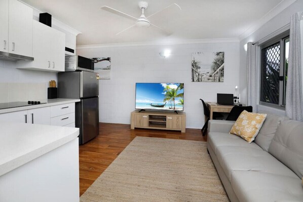 A modern kitchen with white cabinets, a built-in oven, and a refrigerator. There's a flat-screen TV on a wooden stand, a dining area with a table and chairs, and a living room with a sofa and a window.